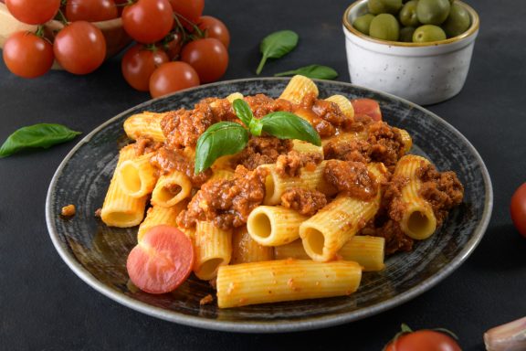 Pasta Pasta mit Bolognese-Soße, Kirschtomaten und frischem Basilikum auf schwarzem Hintergrund.