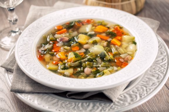 Suppen Gemüsesuppe mit buntem Gemüse in einer weißen Schüssel, serviert auf einem grauen Platz.
