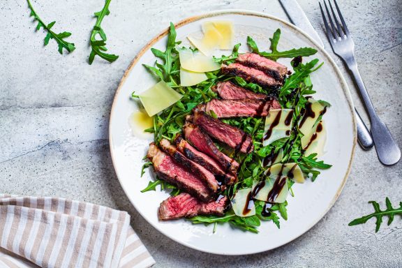 Fleisch Ein Teller mit aufgeschnittenem Steak, Rucola und Parmesan, mit Balsamico-Glasur.